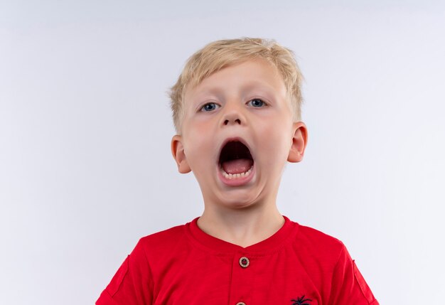 Un pequeño niño rubio lindo con camiseta roja abriendo la boca y gritando mientras mira en una pared blanca