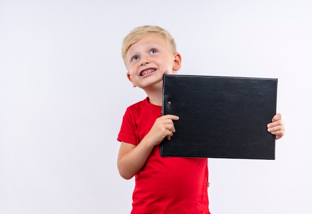 Un pequeño niño rubio lindo en camiseta roja sonriendo y sosteniendo una carpeta en blanco mientras mira hacia arriba en una pared blanca