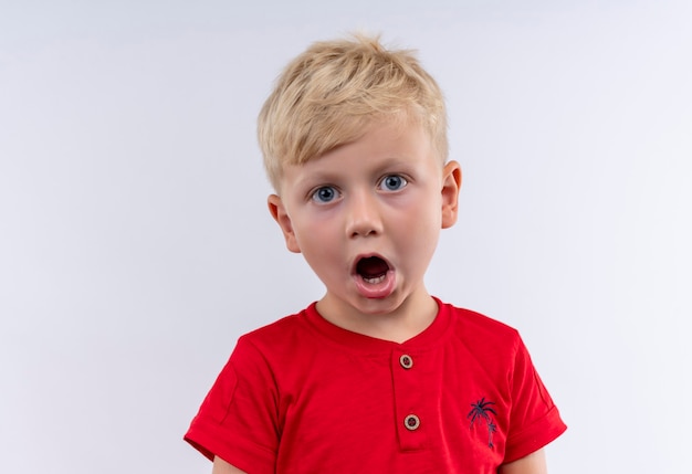 Un pequeño niño rubio lindo en camiseta roja sorprendente mientras mira en una pared blanca