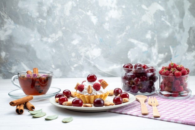 Pequeño pastel cremoso con frambuesas, cerezas y galletas pequeñas, té, canela sobre un escritorio con luz blanca, pastel de frutas, crema de bayas dulces