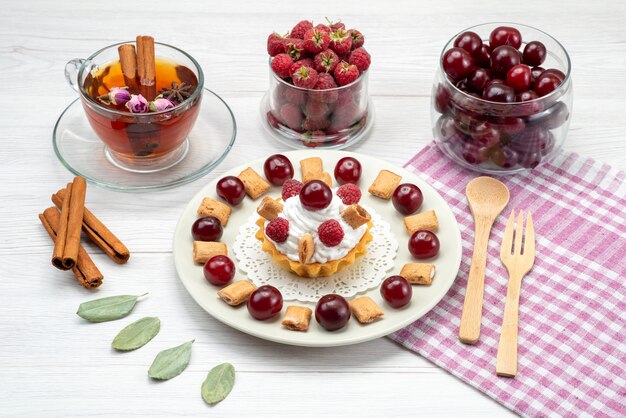 Pequeño pastel cremoso con frambuesas, cerezas y galletas pequeñas, té, canela sobre un escritorio con luz blanca, pastel de frutas, crema de bayas dulces