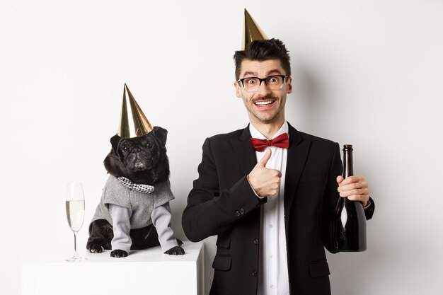 Pequeño perro negro con sombrero de fiesta y de pie cerca de un hombre feliz celebrando las vacaciones, propietario mostrando el pulgar hacia arriba y sosteniendo una botella de champán, fondo blanco.