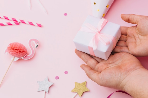 Pequeño regalo en la mano de la persona con accesorios de fiesta sobre fondo rosa