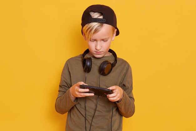 Pequeño tipo concentrado con camisa y gorra casual, jugando videojuegos en línea usando un teléfono móvil, posando con auriculares