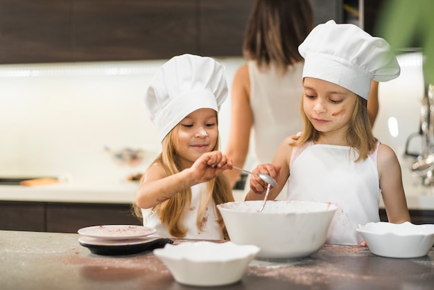 Pequeños hermanos en sombrero de cocinero mezclando ingredientes en un tazón sobre encimera de cocina