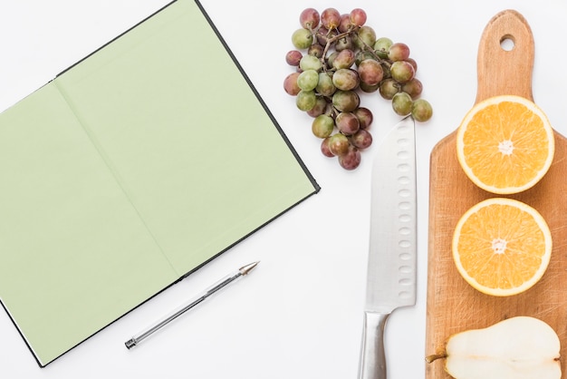 Foto gratuita pera a la mitad frutas de naranja en la tabla de cortar con racimo de uvas; cuchillo; pluma y cuaderno sobre fondo blanco