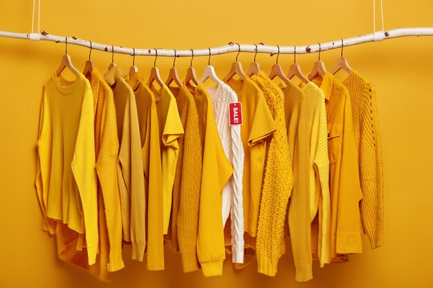 Percha llena de suéteres femeninos de color amarillo sólido. Un suéter blanco destaca de colección, estando en oferta.