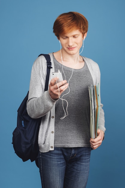 Perder a un joven y atractivo estudiante masculino pelirrojo con un atuendo gris informal con una mochila negra con cuadernos en las manos, usar auriculares, elegir la canción para escuchar en el teléfono inteligente