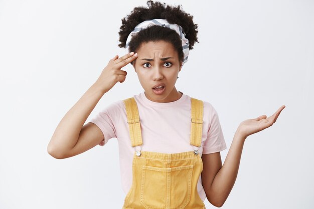 Perdiste la cabeza. Retrato de mujer afroamericana decepcionada confundida y disgustada con diadema y un mono amarillo, sosteniendo el dedo cerca de la sien, encogiéndose de hombros, mirando a un amigo loco o extraño