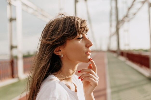 Perfil al aire libre retrato de mujer bonita atractiva en camiseta blanca mirando a otro lado mientras camina en la ciudad en un día cálido y soleado