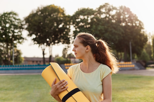 Perfil al aire libre retrato de mujer joven atractiva con camiseta amarilla sosteniendo la estera en las manos, mirando a otro lado, estar listo para hacer ejercicio en el estadio, atención médica.