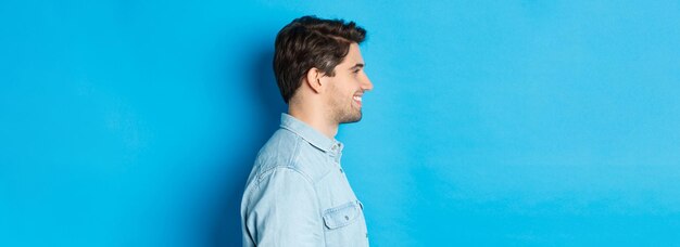 Perfil de un apuesto joven mirando a la izquierda sonriendo feliz de pie sobre fondo azul.