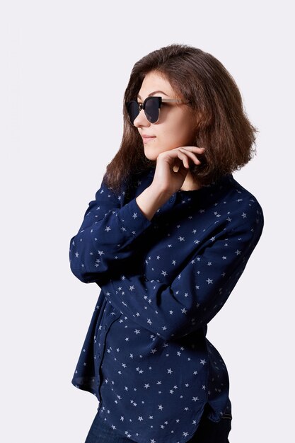 Perfil de elegante mujer morena con peinado de moda vestida con camisa moderna con gafas de sol sosteniendo su mano debajo de la barbilla