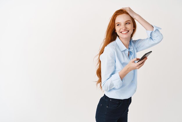 El perfil de una hermosa gerente con cabello largo y pelirrojo usando un teléfono móvil gira la cabeza a la izquierda y sonríe en el espacio de la copia de pie con un teléfono inteligente contra un fondo blanco