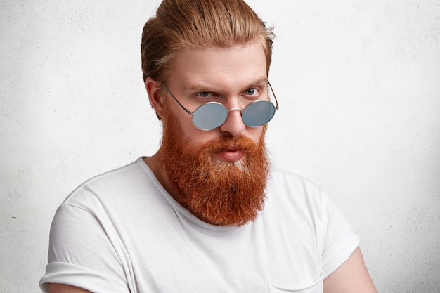 Perfil de hombre barbudo estricto que se ve con confianza a través de gafas de sol, tiene una barba gruesa de jengibre bien recortada y una piel sana, vestido con una camiseta blanca informal