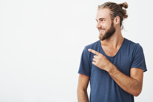 Foto gratuita perfil de hombre guapo alegre con peinado de moda y barba sonriendo brillantemente y apuntando a copyspace