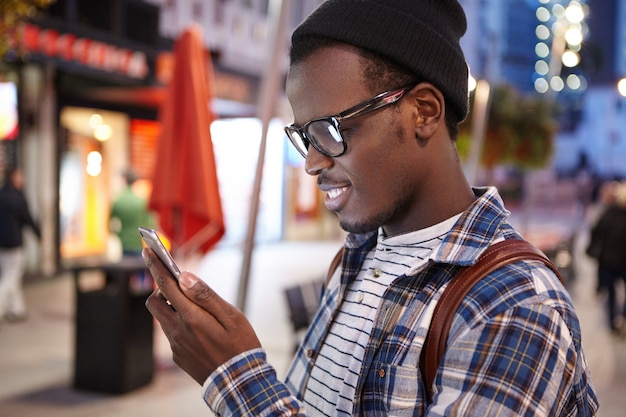 Perfil de un joven turista afroamericano con elegantes gafas y sombrero usando un teléfono inteligente, tratando de encontrar un hostal u hotel para pasar la noche mientras se detiene en otra ciudad extranjera durante su viaje por carretera