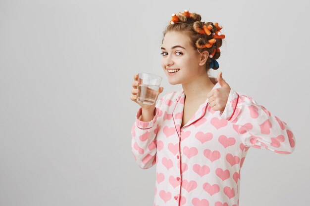 Perfil de mujer joven con rizadores de pelo bebiendo agua en pijama muestran Thumbs-up