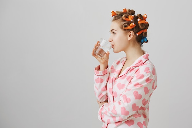 Perfil de mujer joven con rizadores de pelo bebiendo agua en pijama