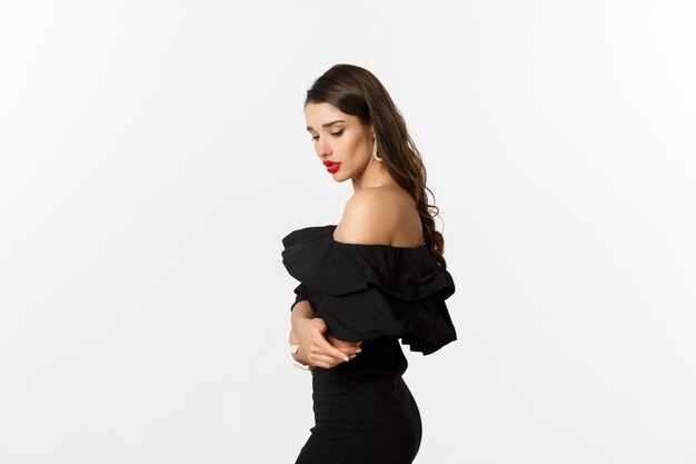 Perfil de mujer morena sensual en elegante vestido negro, mirando hacia abajo, de pie sobre fondo blanco