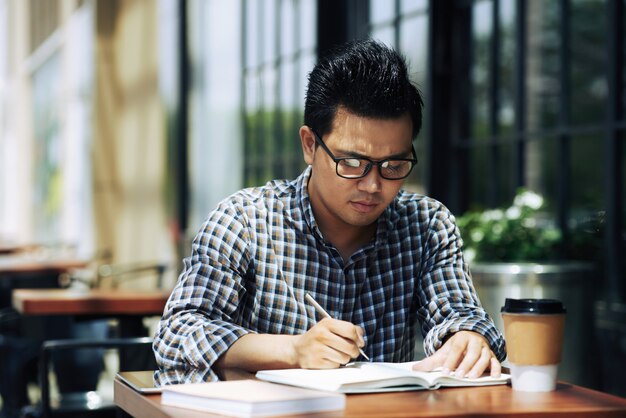 Periodista independiente masculino asiático en gafas sentado en un café al aire libre y escribiendo en el cuaderno