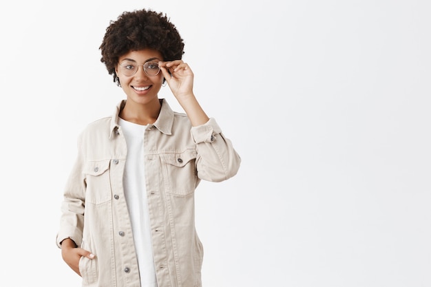 Periodista inteligente y creativa con camisa beige y gafas, tocando el borde de las gafas, sonriendo, sosteniendo la mano en el bolsillo, segura de sí misma y satisfecha con un gran resultado