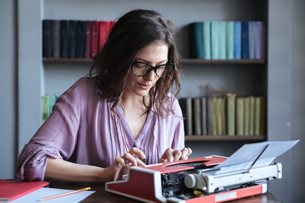 Periodista morena mujer madura en anteojos escribiendo en máquina de escribir en interiores