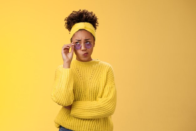 Perplejo inseguro elegante diseñador creativo femenino afroamericano en diadema suéter gafas de sol sonriendo con satisfacción labios plegables tubo incierto tienen dudas frunciendo el ceño mirar hacia arriba pensativo no puede decidir.