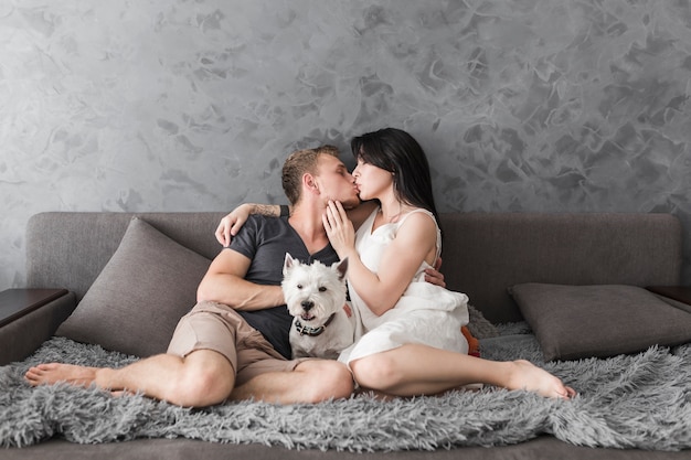 Perro blanco entre la joven pareja sentada en el sofá besándose