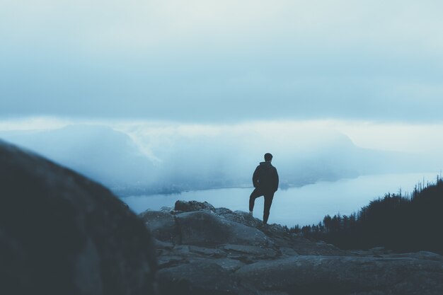 Persona en un abrigo cálido de pie sobre una montaña rocosa y mirando a los árboles