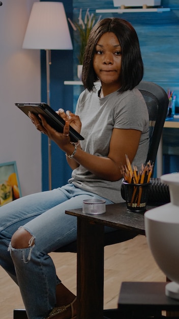 Persona afroamericana con afición artística usando tableta en estudio de taller en casa. Mujer artista negra con tecnología digital trabajando en el dibujo de un jarrón para una obra maestra profesional