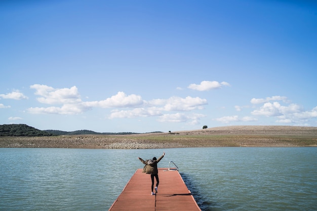 Persona alegre saltando sobre un muelle cerca del lago bajo un cielo nublado