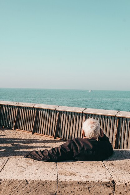 Persona con chaqueta negra sentada en un banco de madera marrón cerca del mar durante el día