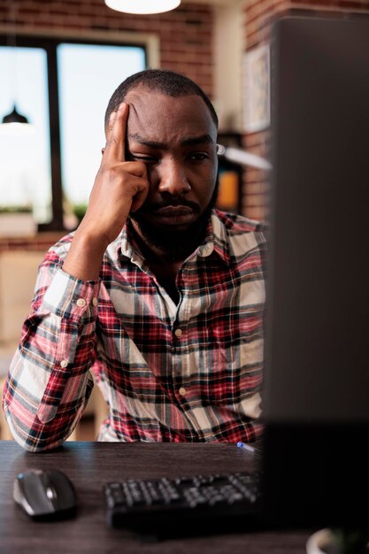 Persona estresada con exceso de trabajo haciendo tareas independientes en la computadora, sintiéndose exhausta y soñolienta en el escritorio de casa. Adulto cansado haciendo trabajo remoto, sintiéndose frustrado por la carga de trabajo de la oficina y los datos de investigación.