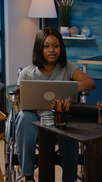 Persona negra no válida con computadora portátil diseñando obras de arte en un espacio creativo en casa. Artística mujer afroamericana en silla de ruedas trabajando con dispositivo en busca de inspiración para obra maestra