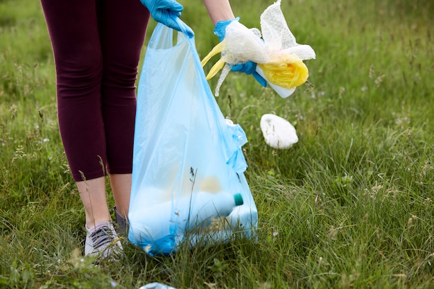 Persona con pantalones de color burdeos recogiendo basura de la hierba verde y tirando la basura en la bolsa del paquete