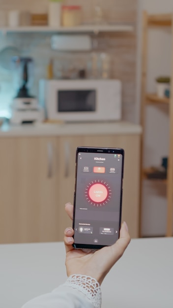 Persona que mira el teléfono móvil con la aplicación de control de iluminación sentado en la cocina de la casa con sistema de luz de automatización, encendiendo la bombilla usando el comando de voz