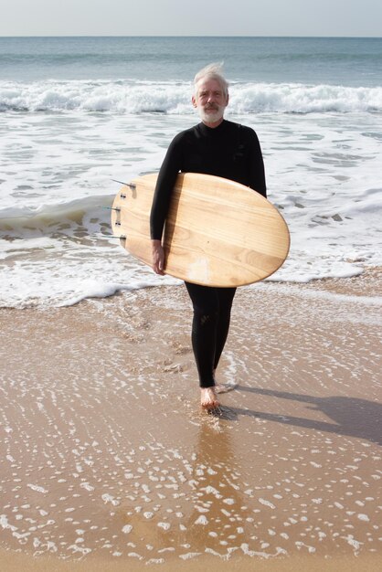 Persona que practica surf mayor que sostiene la tabla de surf grande. Alegre hombre caucásico con traje de neopreno disfrutando del descanso activamente pasando tiempo cuando navega en el mar. Actividad deportiva y concepto de vacaciones de personas mayores.