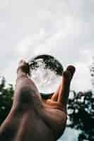 Foto gratuita una persona que sostiene una bola de cristal con el reflejo de hermosos árboles verdes y nubes impresionantes