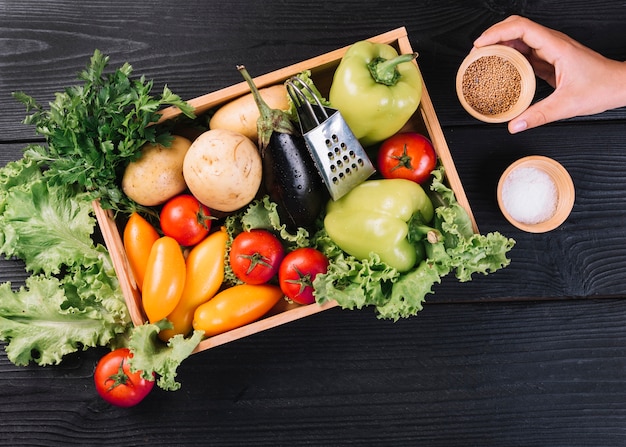 Una persona que sostiene un tazón de mostaza cerca de verduras frescas en un recipiente en una mesa de madera negra