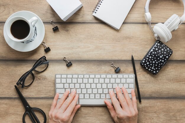 Persona sin rostro escribiendo en el teclado cerca de papelería taza de café y auriculares