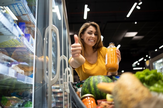 Persona del sexo femenino que sostiene el jugo de naranja en la tienda y mostrando los pulgares para arriba