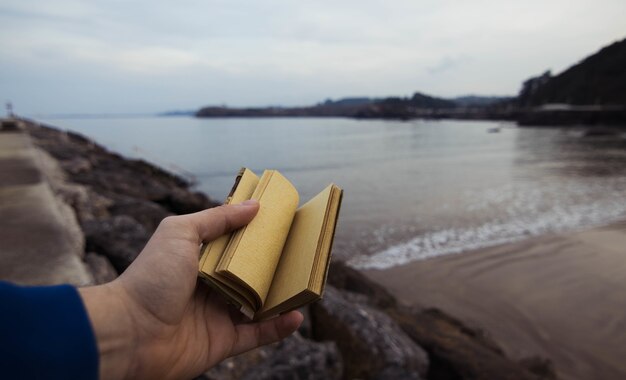 Persona sosteniendo un pequeño cuaderno con un mar borroso en Candás, Asturias, España