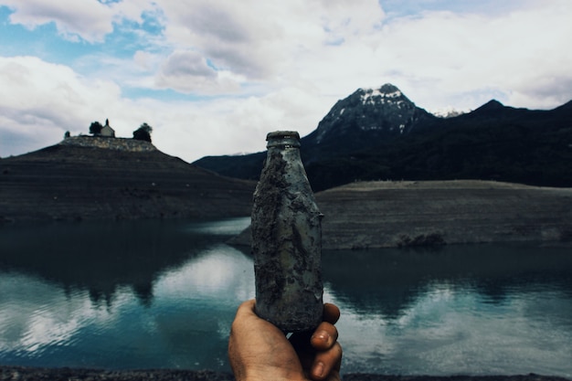Persona sosteniendo una vieja botella de vidrio cubierta de barro cerca del agua con montañas