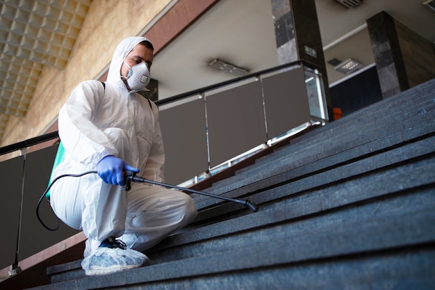 Persona con traje blanco de protección química que desinfecta los pasillos públicos y los pasos para detener la propagación del virus corona altamente contagioso