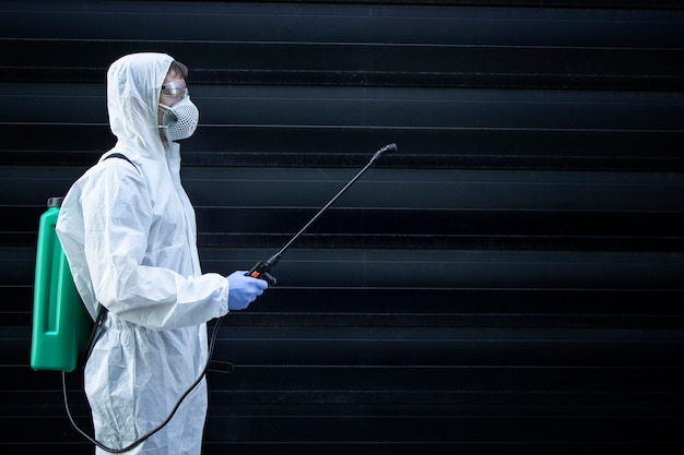 Persona con traje blanco de protección química sosteniendo un rociador con productos químicos desinfectantes para detener la propagación de virus altamente contagiosos