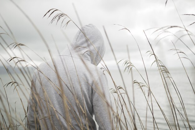 Una persona triste y solitaria por detrás con una sudadera con capucha sentada cerca del mar pensando en la vida.