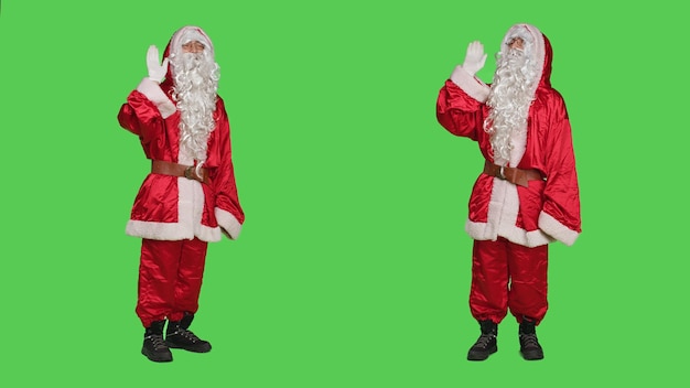 Un personaje amigable saluda y saluda a la gente frente a la cámara, vistiendo trajes festivos tradicionales para difundir el espíritu navideño. Adulto joven vestido como Papá Noel saludando, fondo de pantalla verde.