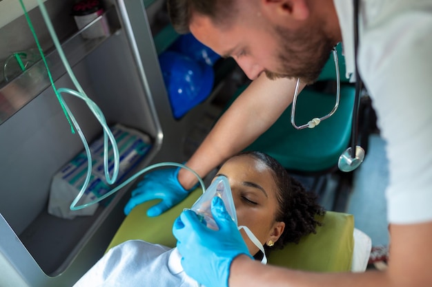 Personal de emergencia de atención médica paramédico que cuida a una paciente joven acostada en camilla con sistema de ventilación médica y máscaras de válvula de bolsa dentro de un camión de ambulancia de servicio médico