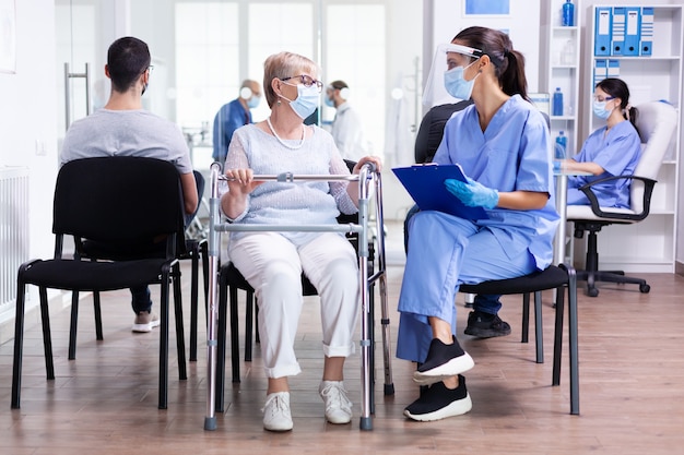 Personal médico discutiendo el tratamiento con una mujer mayor discapacitada que lleva un andador en la sala de espera del hospital con mascarilla contra el coronavirus